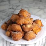 Side view of Bolinhos de Chuva (Donut holes with Cinnamon Sugar coating) stacked on a plate in a pyramid with 1 donut on top. Plate is on the marble counter with blue and white striped towel under.
