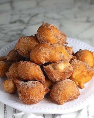 Side view of Bolinhos de Chuva (Donut holes with Cinnamon Sugar coating) stacked on a plate in a pyramid with 1 donut on top. Plate is on the marble counter with blue and white striped towel under.