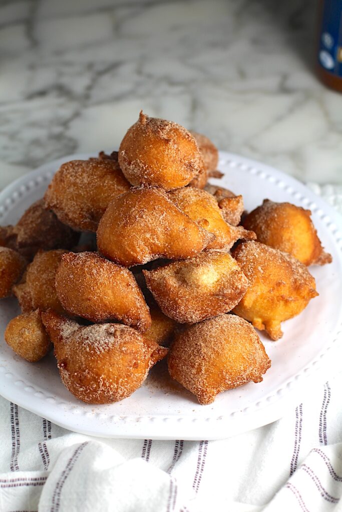 Side view of Bolinhos de Chuva (Donut holes with Cinnamon Sugar coating) stacked on a plate in a pyramid with 1 donut on top.  Plate is on the marble counter with blue and white striped towel under.