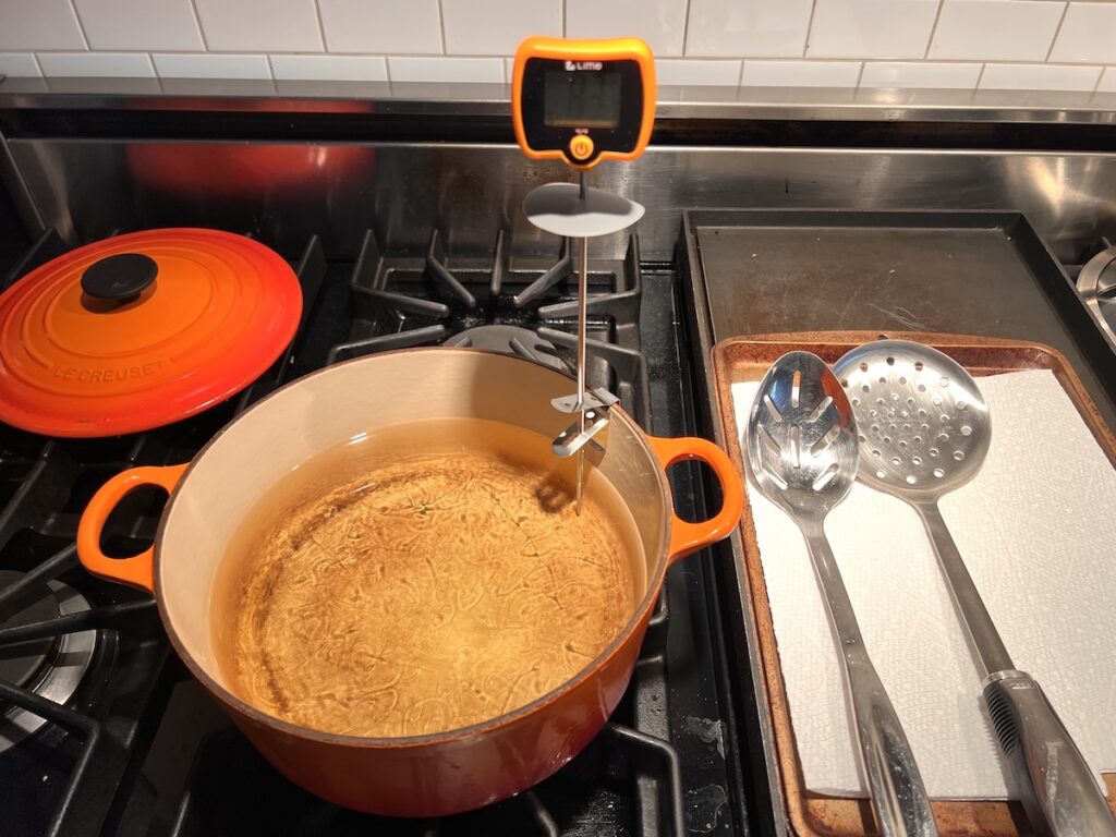 Everything set up for frying the Bolinhos de Chuva with a pot containing oil, a thermometer attached to the pot, a sheet pan at the side lined with paper towels and a slotted spoons.